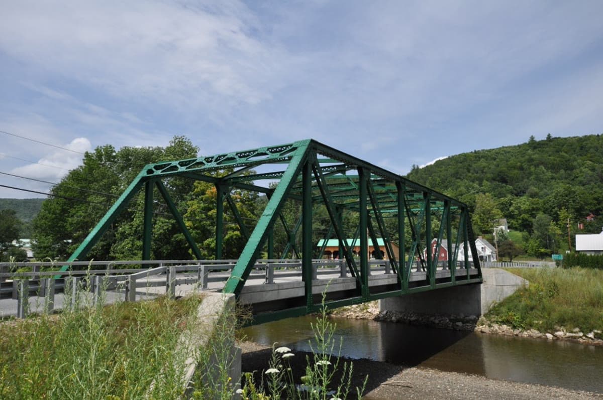A green truss bridge over the Ottauquechee River in Bridgewater Corners, Vermont with a red barn and forested hills